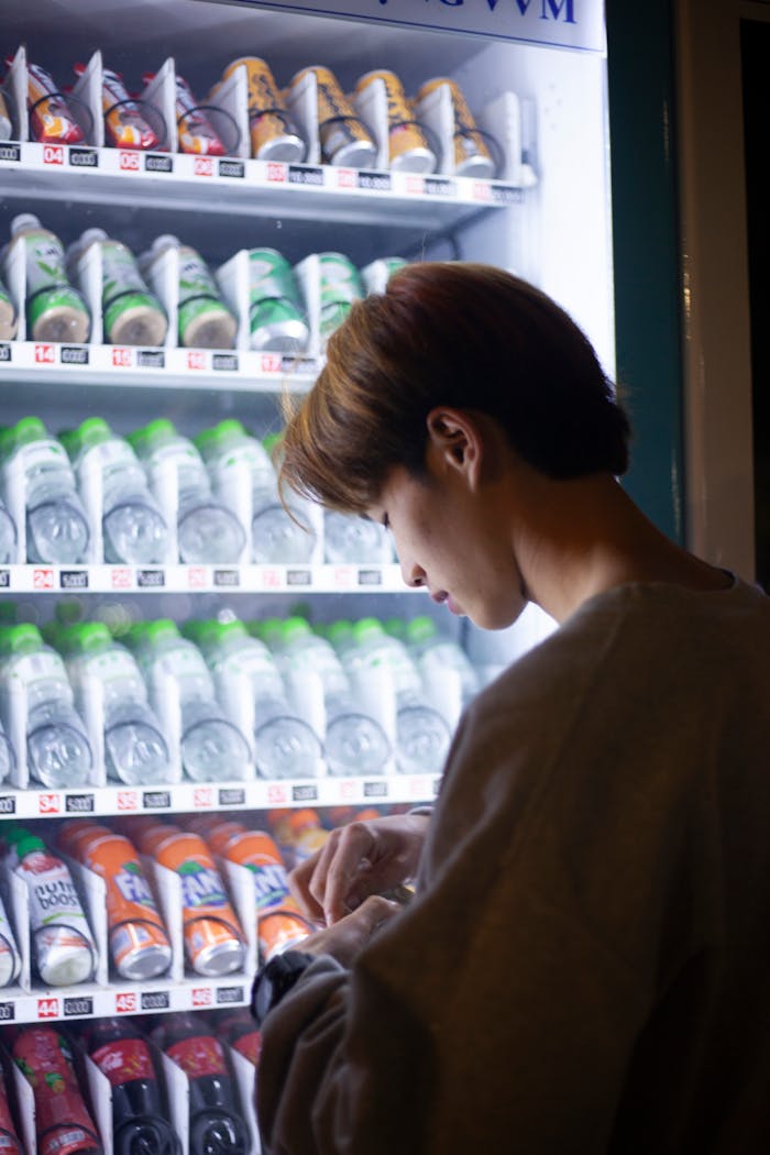 A man standing at a vending machine, selecting a drink under soft lighting.